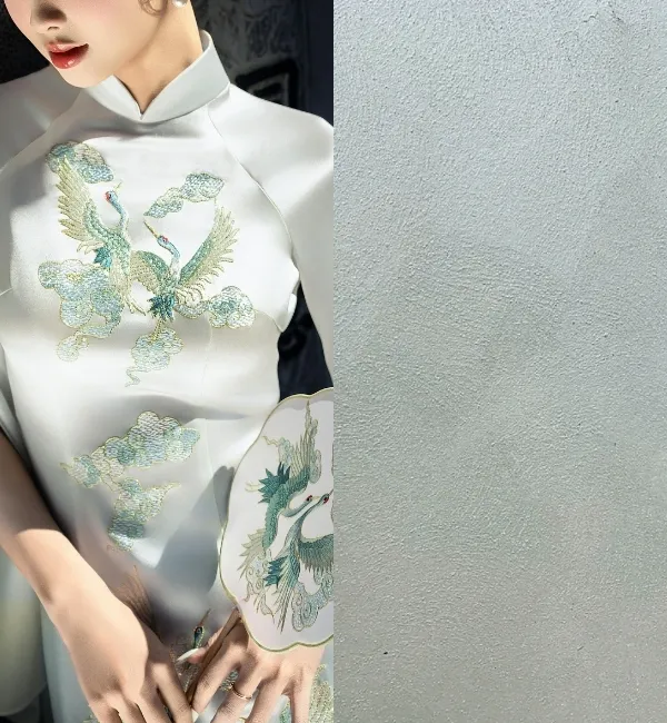 A woman wears a white ao dai with embroidered cranes and clouds, holding a fan.