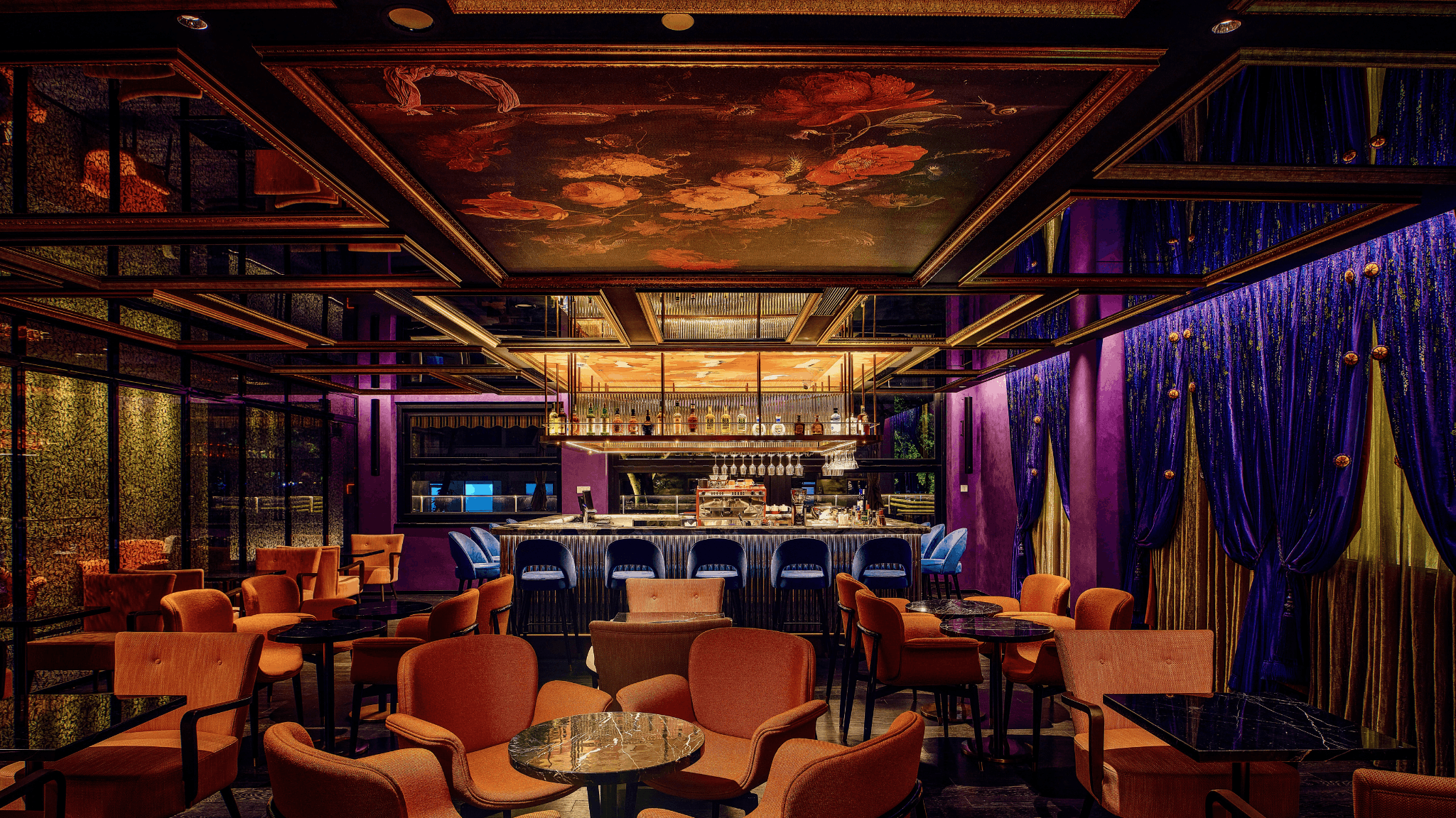 Dramatic lounge bar interior with floral-painted ceiling panels, purple velvet drapes, orange upholstered armchairs