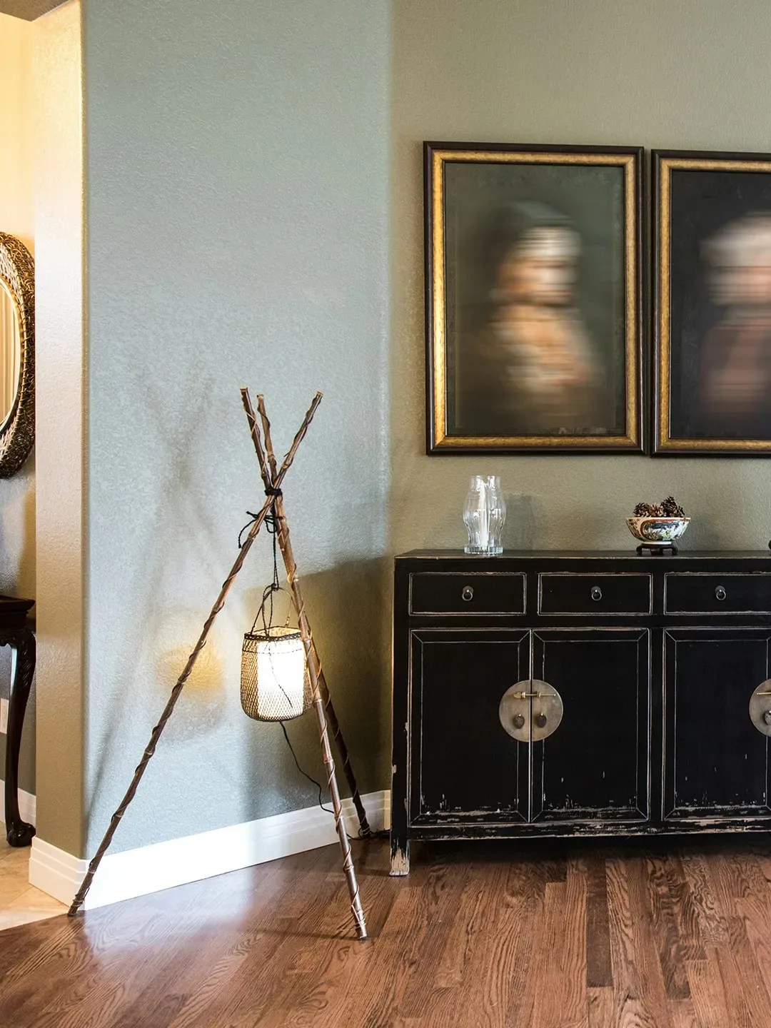 A rustic tripod lamp with a caged bulb stands beside a distressed black cabinet with ornate hardware.