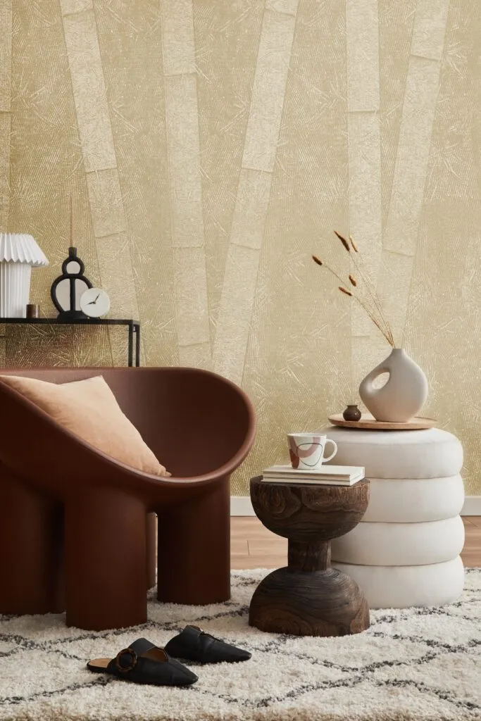 A modern living room features a brown chair, white poufs, and a textured gold wallpaper.