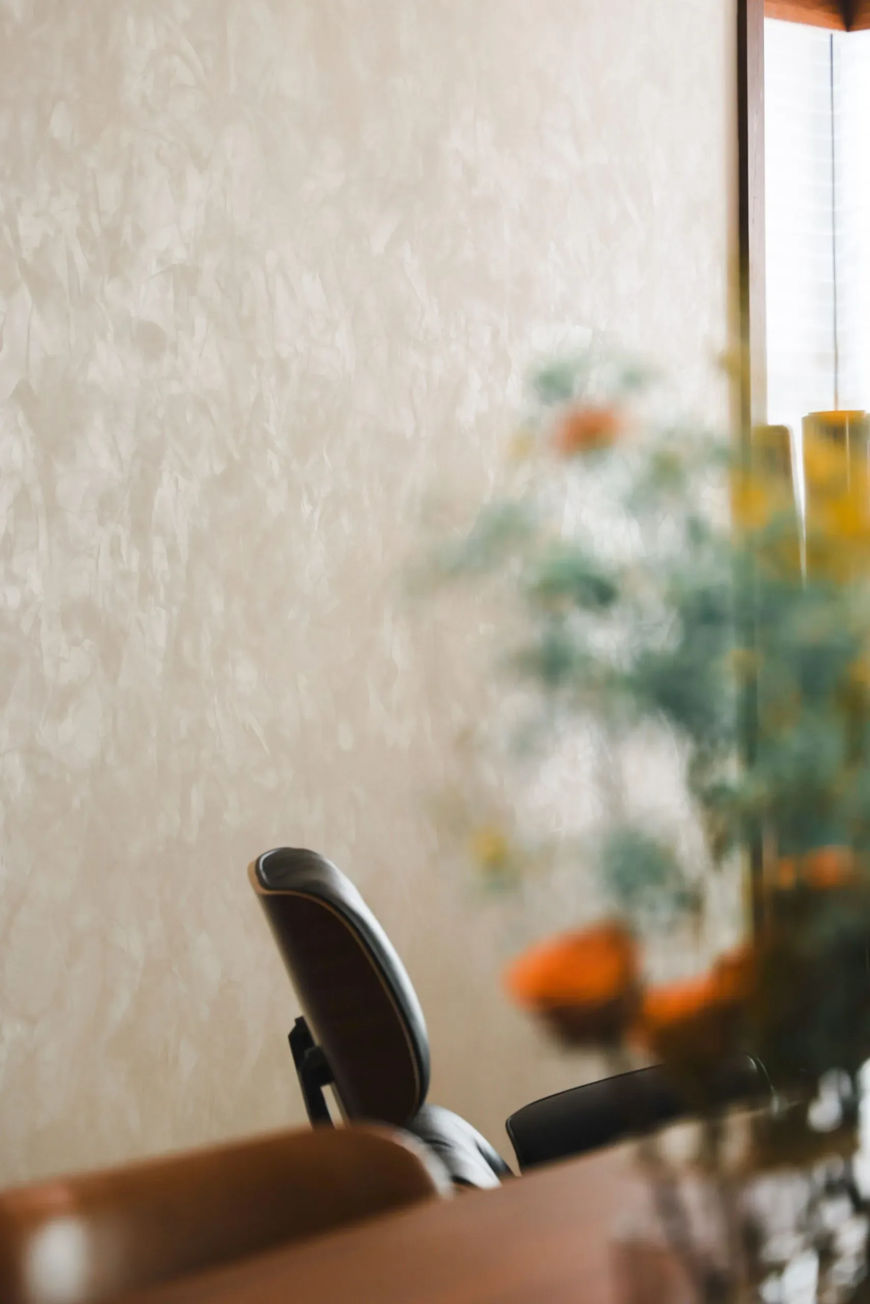 A textured cream wall behind a dark leather chair and blurred greenery with orange accents.