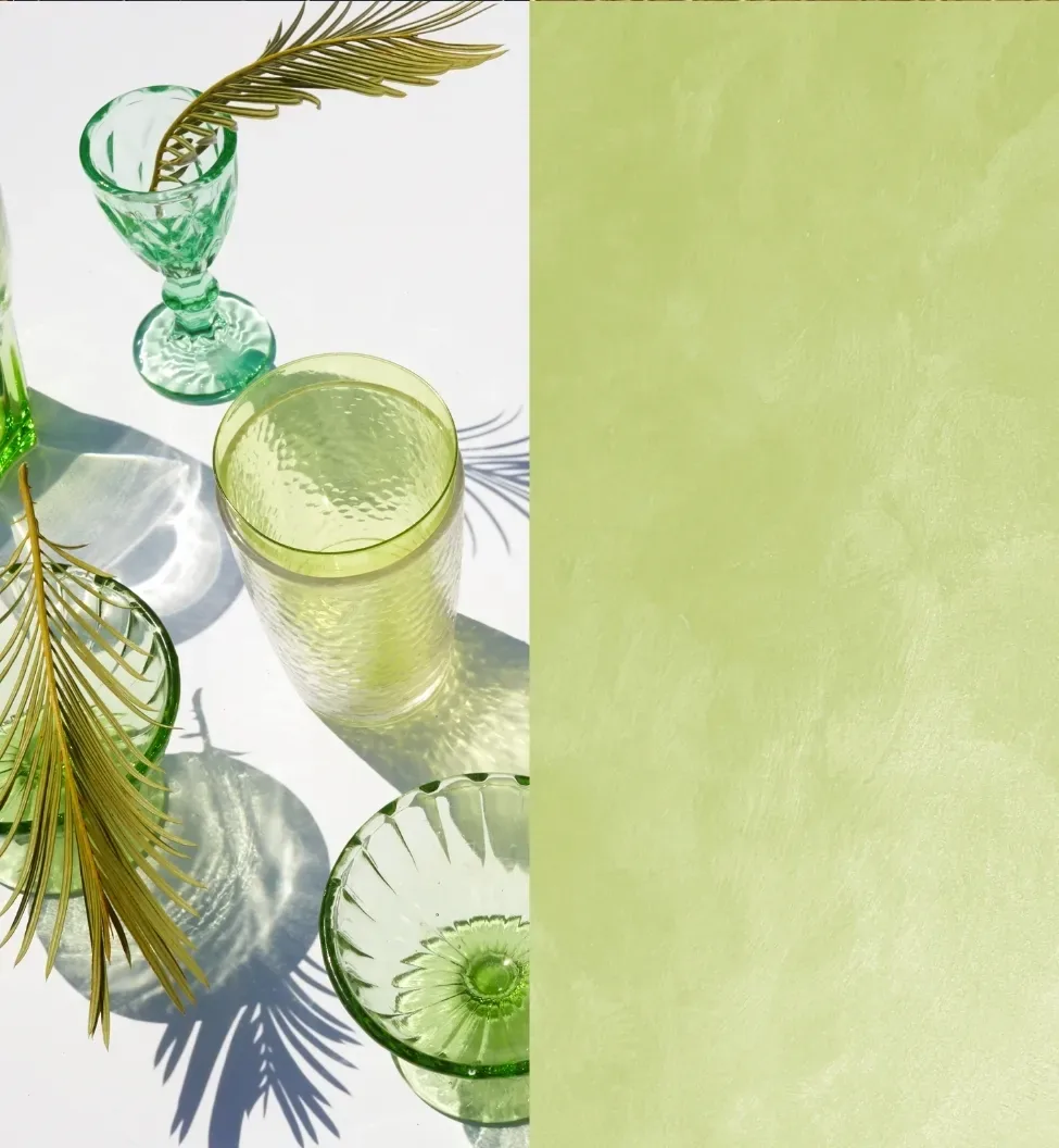 Green glassware and palm fronds cast shadows on a white surface next to a textured green background.