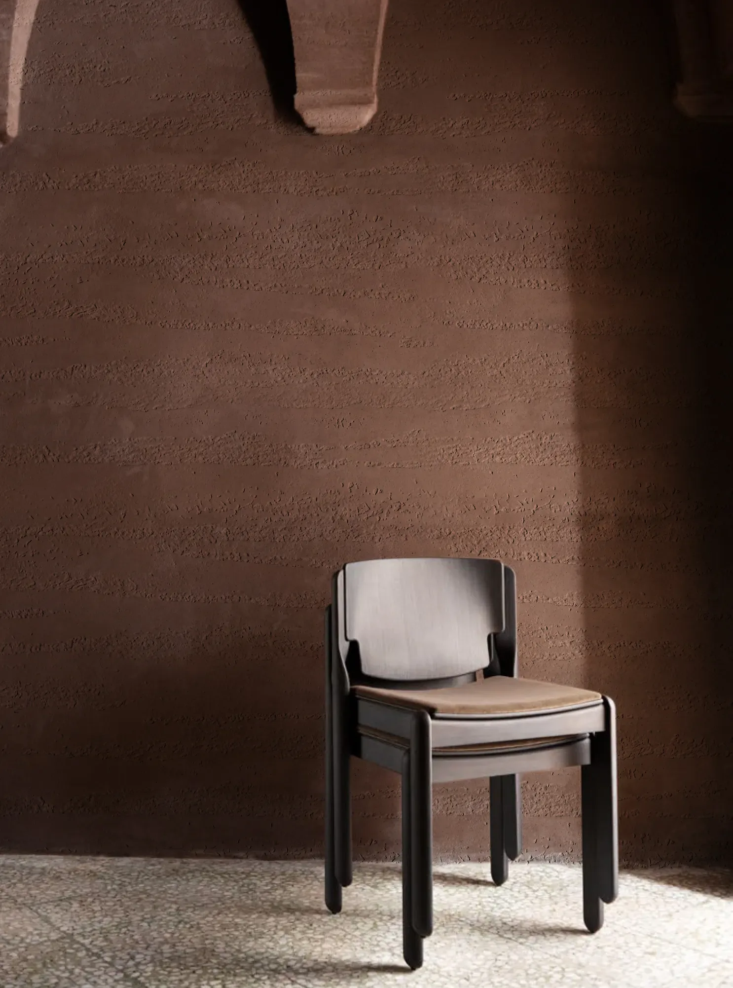 A modern dark wood chair with a tan seat rests on a speckled floor against a textured brown wall.