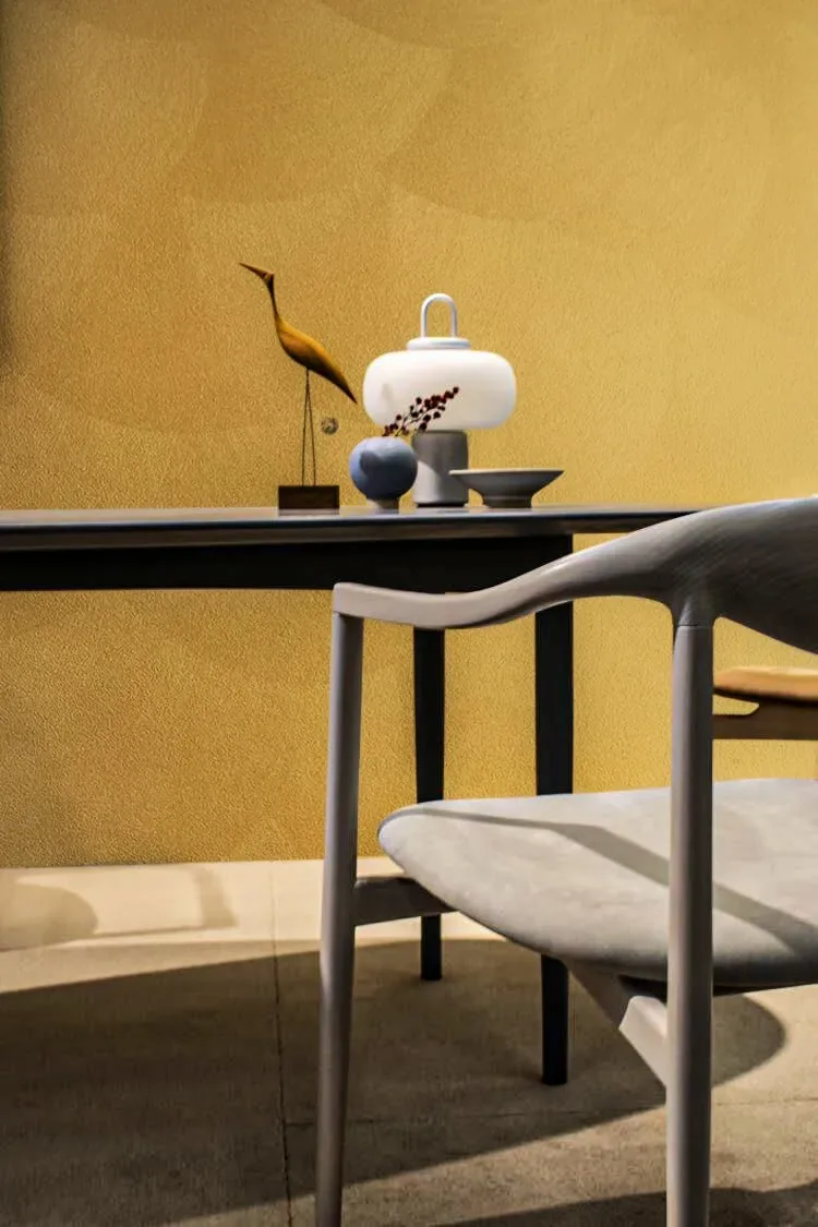 A modern gray chair sits before a dark table with decorative objects against a textured yellow wall.