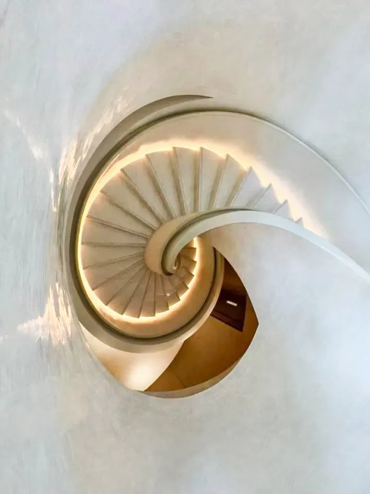 A modern spiral staircase with warm lighting curves elegantly within a textured white architectural space.
