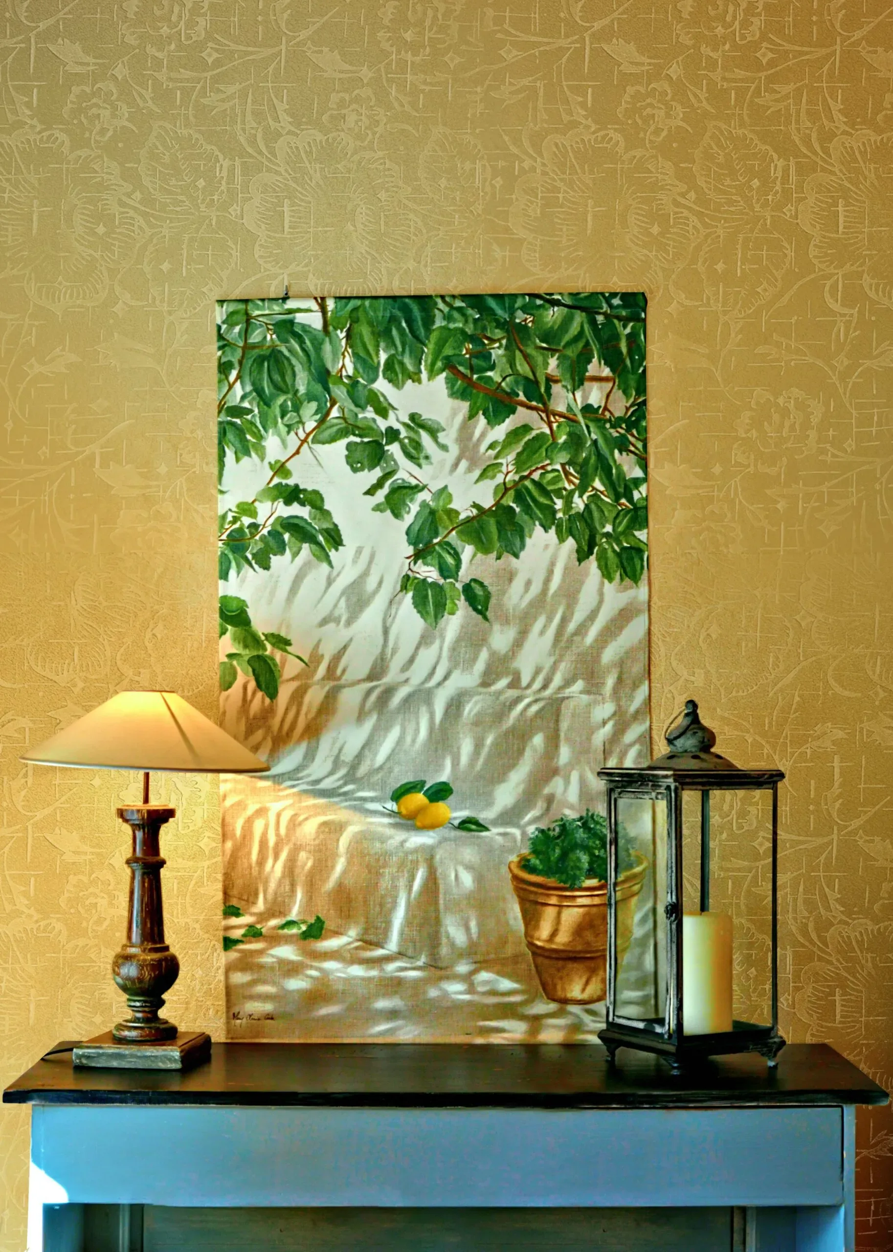 A still life painting of lemons and greenery hangs above a table with a lamp and lantern.