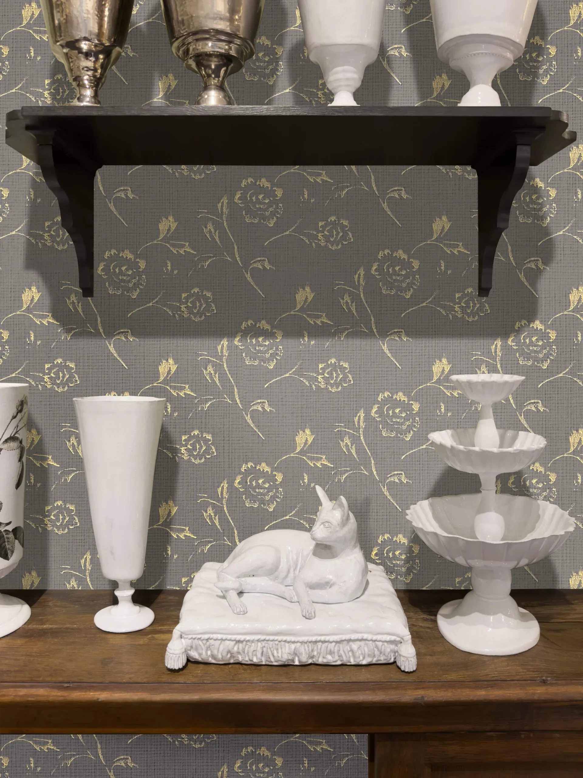 A white ceramic cat rests on a cushion next to a tall vase on a wooden shelf.