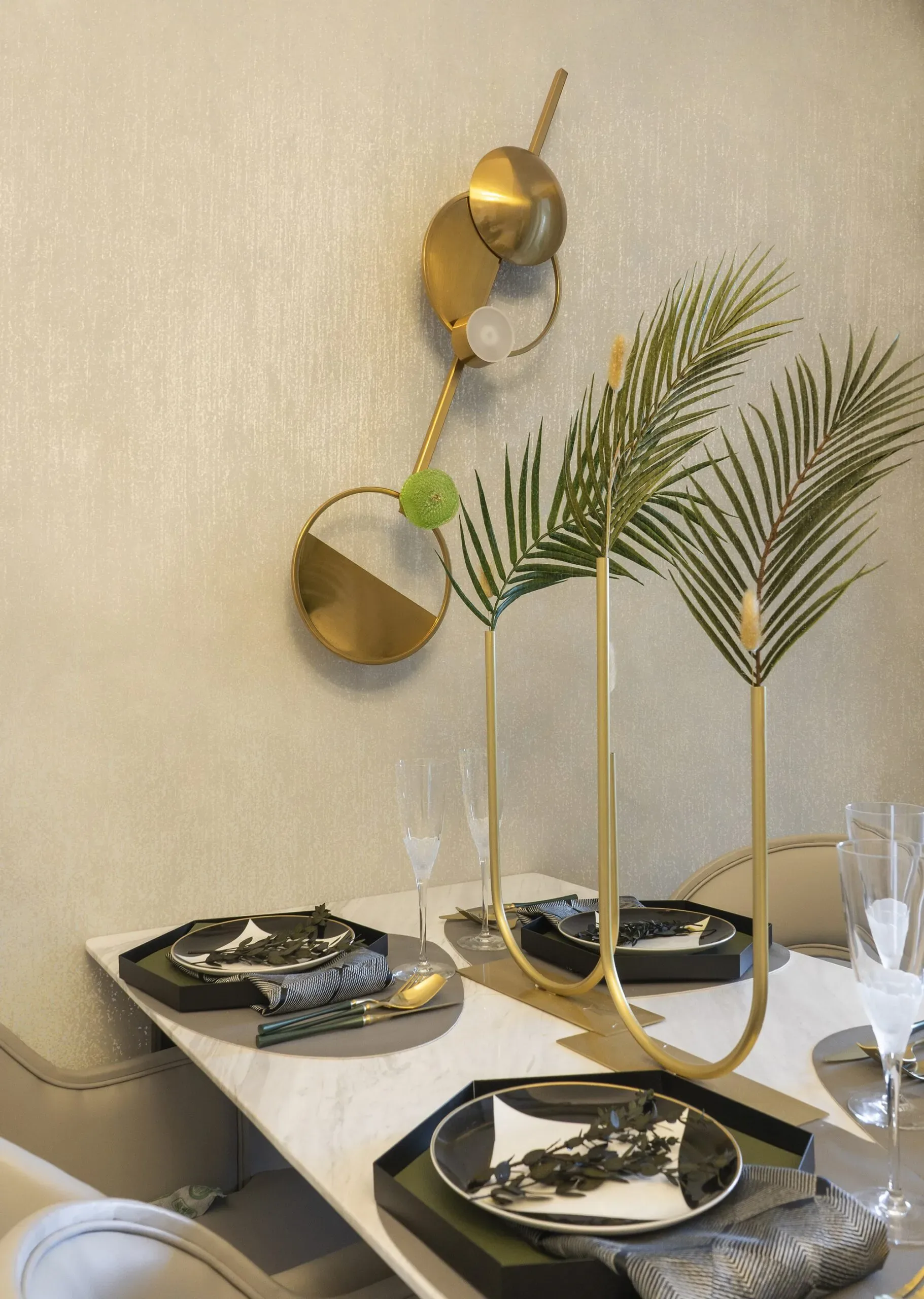 A modern dining table is set with black plates, gold cutlery, and palm fronds in a gold vase.