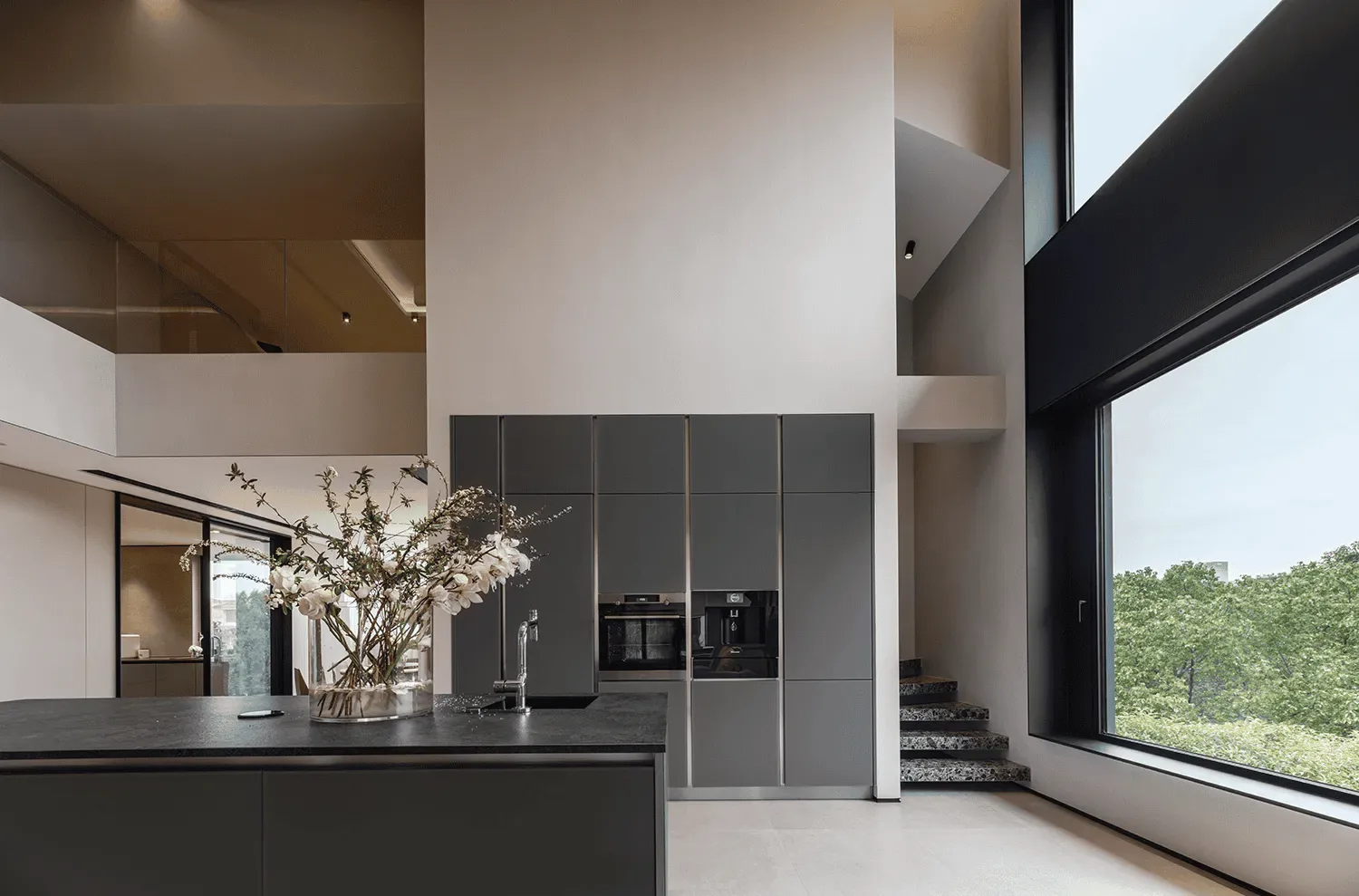 A modern kitchen island with a floral arrangement sits before sleek gray cabinets and a large window.